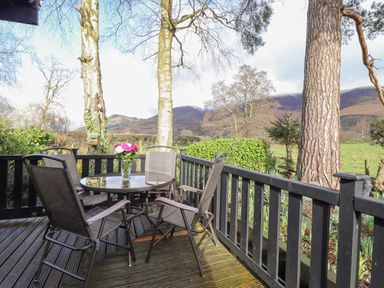 An outdoor deck with a glass table and four chairs with trees and hills in the background at Hayeswater Lodge Burnside Park near Keswick