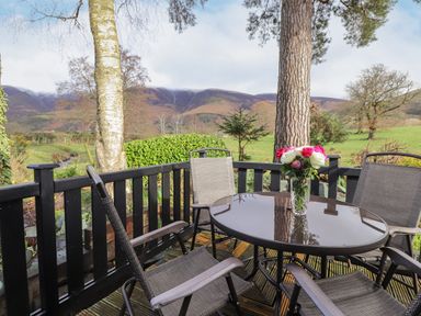 An outdoor patio with a round glass table and four chairs with flowers on the table and trees and hills in the background at Hayeswater Lodge Burnside Park near Keswick