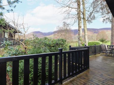 A wooden deck with black railing chairs and a table overlooking bushes trees and distant hills at Hayeswater Lodge Burnside Park near Keswick