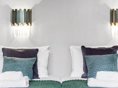Two beds with white sheets and pillows each with a dark pillow and a green patterned cushion under wall lights at Tudor Grange in Bournemouth