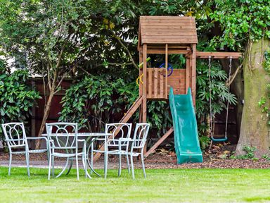 A backyard with a wooden playset including a slide and swing and a metal table with four chairs on grass at Tudor Grange in Bournemouth