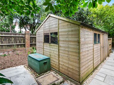 A wooden garden shed next to a green storage box on a stone paved path surrounded by trees and a wooden fence at Tudor Grange in Bournemouth