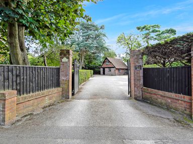 An open driveway gate with brick pillars leading to a parking area and a small brick building surrounded by trees at Tudor Grange in Bournemouth