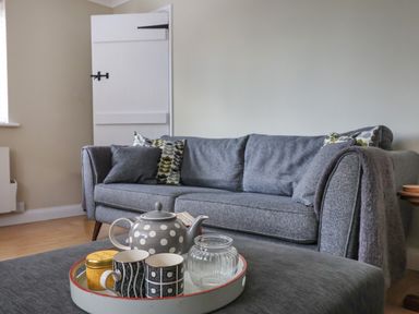 A grey sofa with patterned cushions and throw in a living room with a tray containing a teapot and mugs on a grey ottoman at 8 Biddlecombe Orchard in Bridport