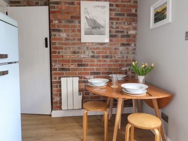 A small dining area with a wooden table and stools against a brick wall with framed pictures and a vase of yellow flowers at 8 Biddlecombe Orchard Bridport