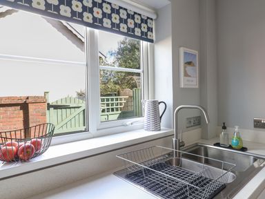 A kitchen sink area with a dish rack a wire fruit basket with apples and a window with floral blinds at 8 Biddlecombe Orchard in Bridport