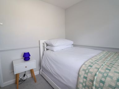A bedroom with a single bed white bedding pillows and a patterned blanket next to a white bedside table with a blue lamp at 8 Biddlecombe Orchard Bridport