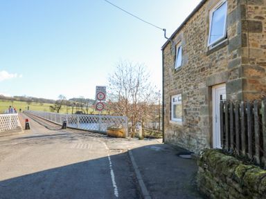 A narrow road leading to a metal bridge next to a stone house with windows and a door at Bridge End Cottage in Wark