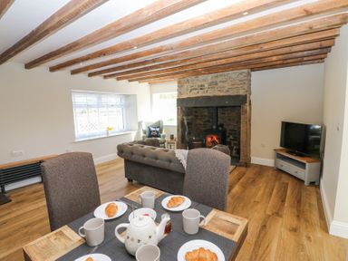 A living room with a wooden beamed ceiling a stone fireplace with wood stove a television a dining table set with tea and croissants and a window with blinds at Bridge End Cottage in Wark