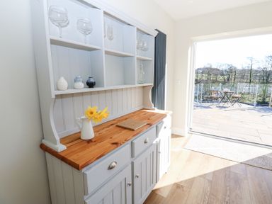A wooden cabinet with glassware pottery and a pitcher with flowers next to a sliding door leading to a patio at Bridge End Cottage in Wark