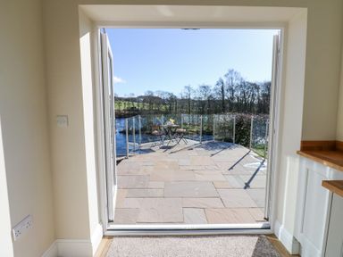 An open doorway leading to a stone patio with a table and two chairs overlooking trees and a river at Bridge End Cottage in Wark