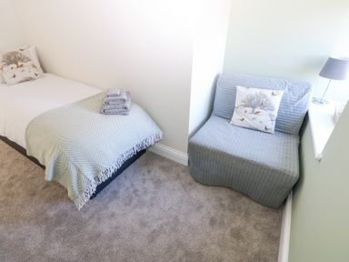 A bedroom with a single bed covered with a blanket and folded towels next to a small sofa with a cushion and a lamp on a white windowsill at Bridge End Cottage in Wark