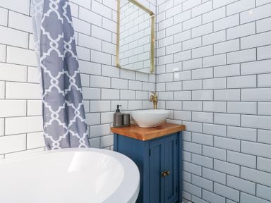 A bathroom corner with a white bathtub a patterned shower curtain a blue cabinet with a wooden countertop and a white vessel sink with a brass faucet and a mirror on tiled walls at Bridge End Cottage in Wark