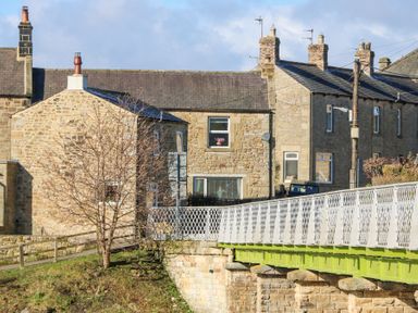 A stone building next to a bridge with green supports and white railings at Bridge End Cottage in Wark