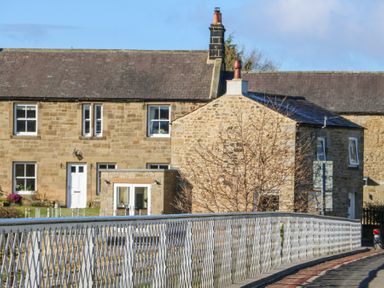 Stone houses behind a metal railing fence and a road at Bridge End Cottage in Wark