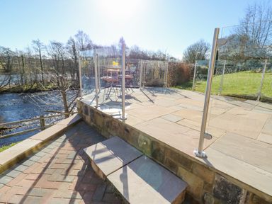 An outdoor patio with glass and metal railings overlooking a river with a table and chairs on the patio at Bridge End Cottage in Wark