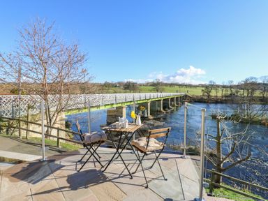 An outdoor seating area with two chairs and a table overlooking a river and a bridge at Bridge End Cottage in Wark