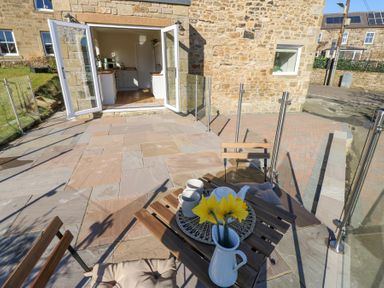 An outdoor patio with wooden chairs and table set with cups teapot and flowers outside a stone building at Bridge End Cottage in Wark