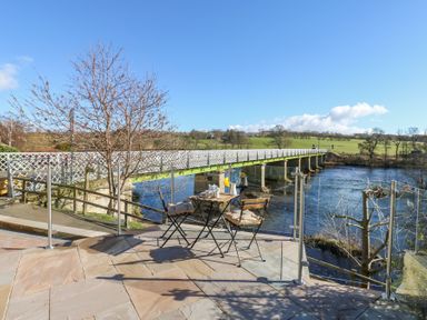An outdoor patio with a small table and two chairs overlooking a river and a bridge at Bridge End Cottage in Wark