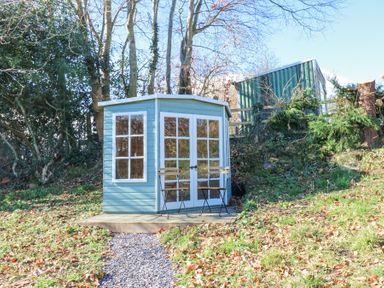 A small blue wooden shed with glass windows and double doors with two chairs outside on a gravel path surrounded by trees at Bridge End Cottage in Wark