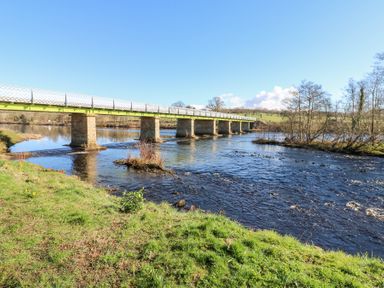 A river with a long bridge supported by stone pillars and trees along the bank at Bridge End Cottage in Wark