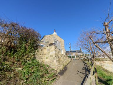 A stone house with a chimney at the top of a paved pathway with stone walls on each side and leafless trees at Bridge End Cottage in Wark