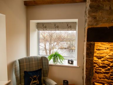 An armchair with a bee cushion next to a window with a plant and clock on the sill and a stone fireplace at Bridge End Cottage in Wark