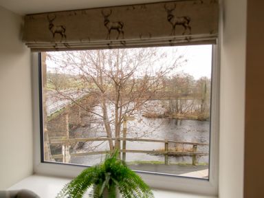 A window with a deer patterned blind overlooking a river with a leafless tree and a bridge at Bridge End Cottage in Wark