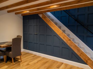 A dining area with two chairs and a table next to a staircase with blue paneled walls and exposed wooden beams at Bridge End Cottage in Wark