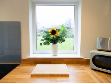 A kitchen counter with a cutting board a microwave a toaster and a vase with a sunflower in front of a window at Bridge End Cottage in Wark
