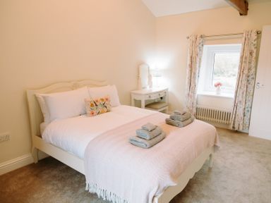 A bedroom with a bed with white linens and folded towels a dressing table with a lamp and a window with floral curtains at Bridge End Cottage in Wark