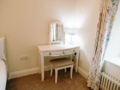 A white dressing table with three drawers a small mirror a stool and a lamp with floral curtains and a radiator at Bridge End Cottage in Wark