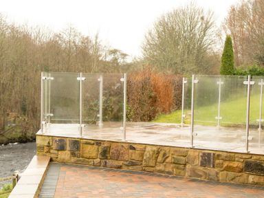 A stone patio with glass railings overlooking a river and trees at Bridge End Cottage in Wark