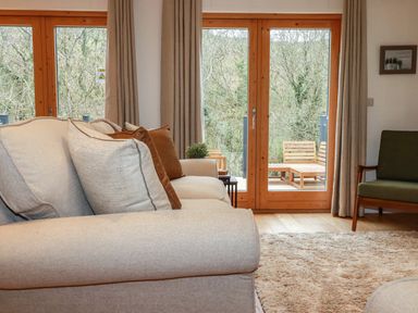 A living room with a beige sofa and cushions a green armchair wooden framed glass doors leading to an outdoor seating area at 1 Streamside in Lanreath