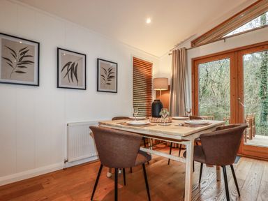 A dining area with a wooden table and four chairs near large glass doors and three framed botanical prints on the wall at 1 Streamside in Lanreath