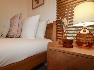 A bed with pillows next to a wooden bedside table with a lamp and decorative items at 1 Streamside in Lanreath