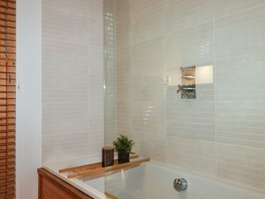 A bathroom with a bathtub with a wooden tray holding a plant and a candle and tiled walls at 1 Streamside in Lanreath
