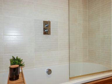 A bathtub with a wooden tray holding a plant and container in a tiled bathroom at 1 Streamside in Lanreath