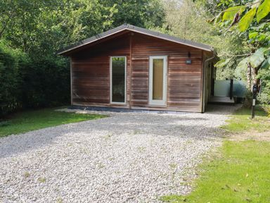 A wooden cabin with a gravel driveway and surrounded by trees at 1 Streamside in Lanreath