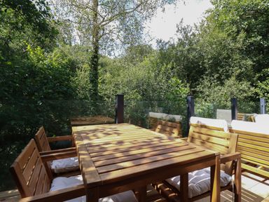 An outdoor wooden dining table with chairs and white cushions on a balcony surrounded by green trees at 1 Streamside in Lanreath
