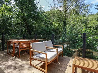 An outdoor wooden deck with chairs a table and glass railing surrounded by trees at 1 Streamside in Lanreath
