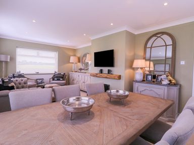 A dining room with a wooden table and silver bowls and a living area with armchairs a television and lamps at The Farmhouse in St Osyth