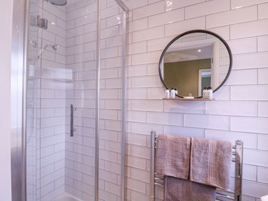 A bathroom with a glass shower enclosure a round mirror above a shelf and towels on a heated rack at The Farmhouse in St Osyth
