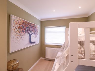 A small bedroom with a white bunk bed a window with blinds a radiator a large colorful tree painting and a lamp at The Farmhouse in St Osyth