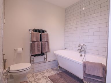 A bathroom with a white toilet a white clawfoot bathtub with towels on the side a towel rack with towels and patterned floor tiles at The Farmhouse in St Osyth