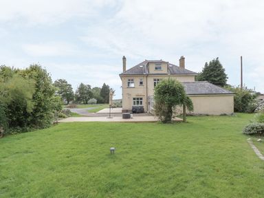 A backyard with a large green lawn and a beige two-story house with a wooden deck at Tan Y Fron in Dyffryn Ardudwy