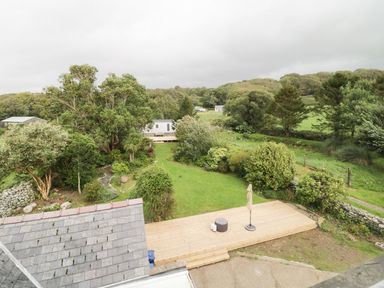A garden with trees bushes a lawn wooden deck with umbrella and stool at Tan Y Fron in Dyffryn Ardudwy
