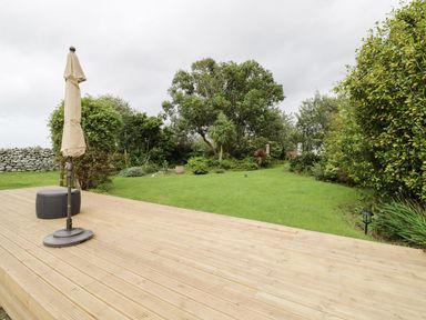 A wooden deck with a closed umbrella and a small round ottoman overlooking a green garden with various trees and bushes at Tan Y Fron in Dyffryn Ardudwy