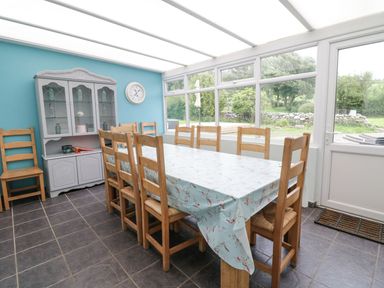 A dining area with wooden chairs and a table covered with a floral tablecloth near large windows and a glass door at Tan Y Fron in Dyffryn Ardudwy