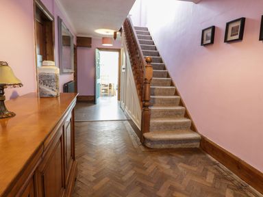 A hallway with wooden floor a carpeted staircase wooden cabinet with a lamp and vase and a door leading to a dining area at Tan Y Fron in Dyffryn Ardudwy
